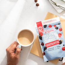 Vegums wholefruit probiotic gummy packet with womans hand holding cup of cappuccino blue nails and two of the gummies on marble background as well as a pair of glasses situated to the right top corner. 