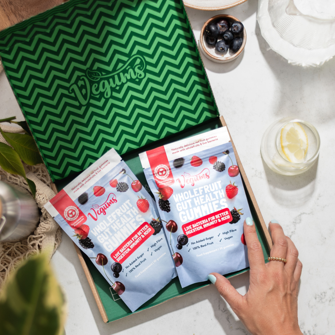 Vegums wholefruit gut health gummies in an eco friendly cardboard delivery box with womans hand holding the edge. There is a glass of water with lemon inside as well as blueberries in a bowl on the marble background.