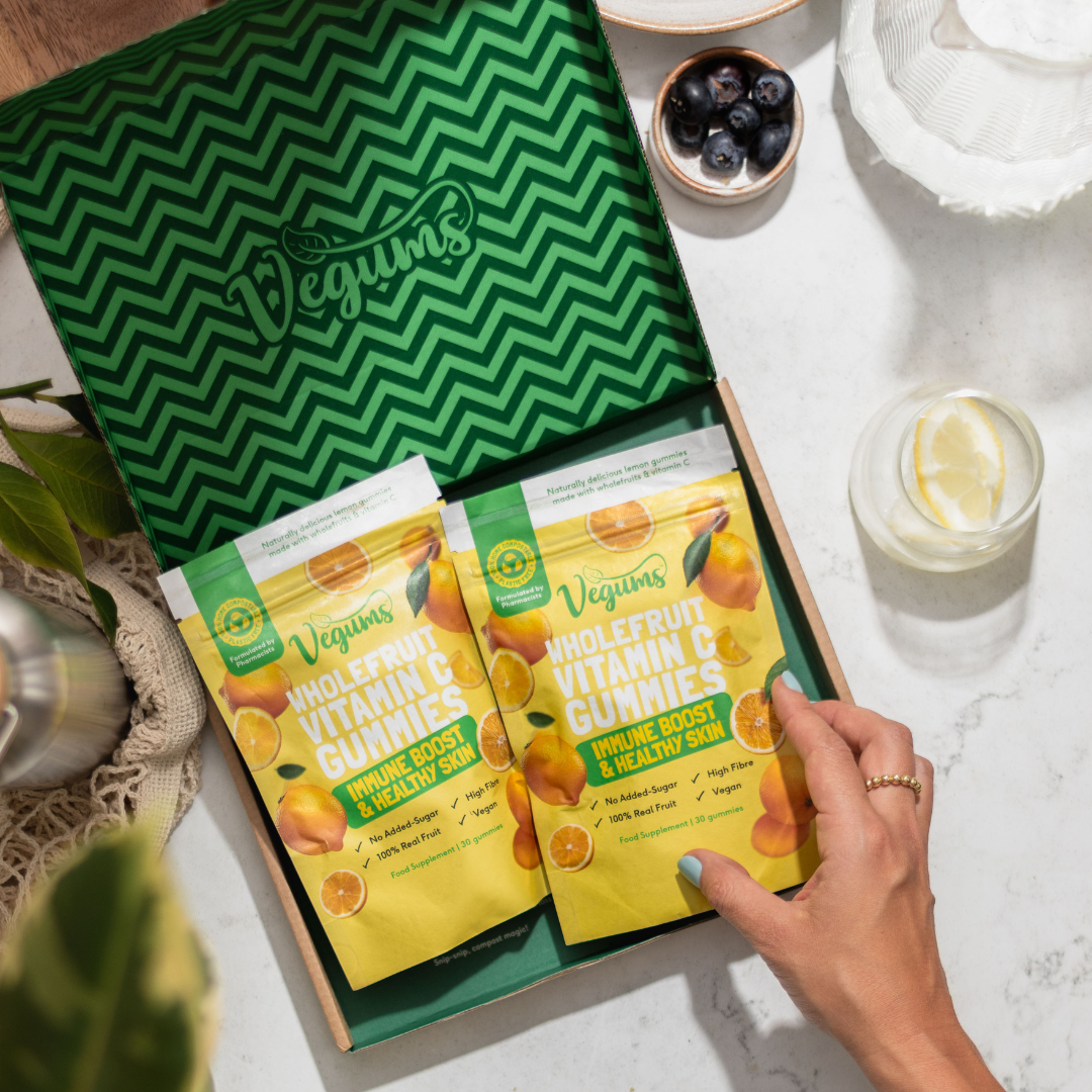 Vegums wholefruit vitamin c gummies in a eco friendly cardboard box woman holding edge of item with a lemon water and berries on a marble background