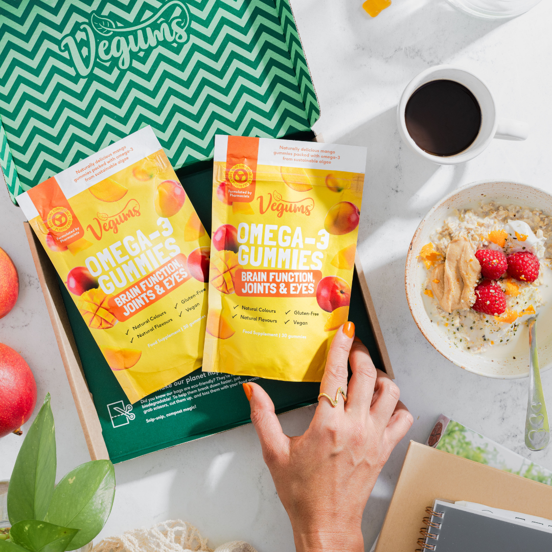 Vegums vegan omega 3 ackets inside eco friendly box woman holding packet with orange nails, marble background with cup of coffee and cereal bowl 